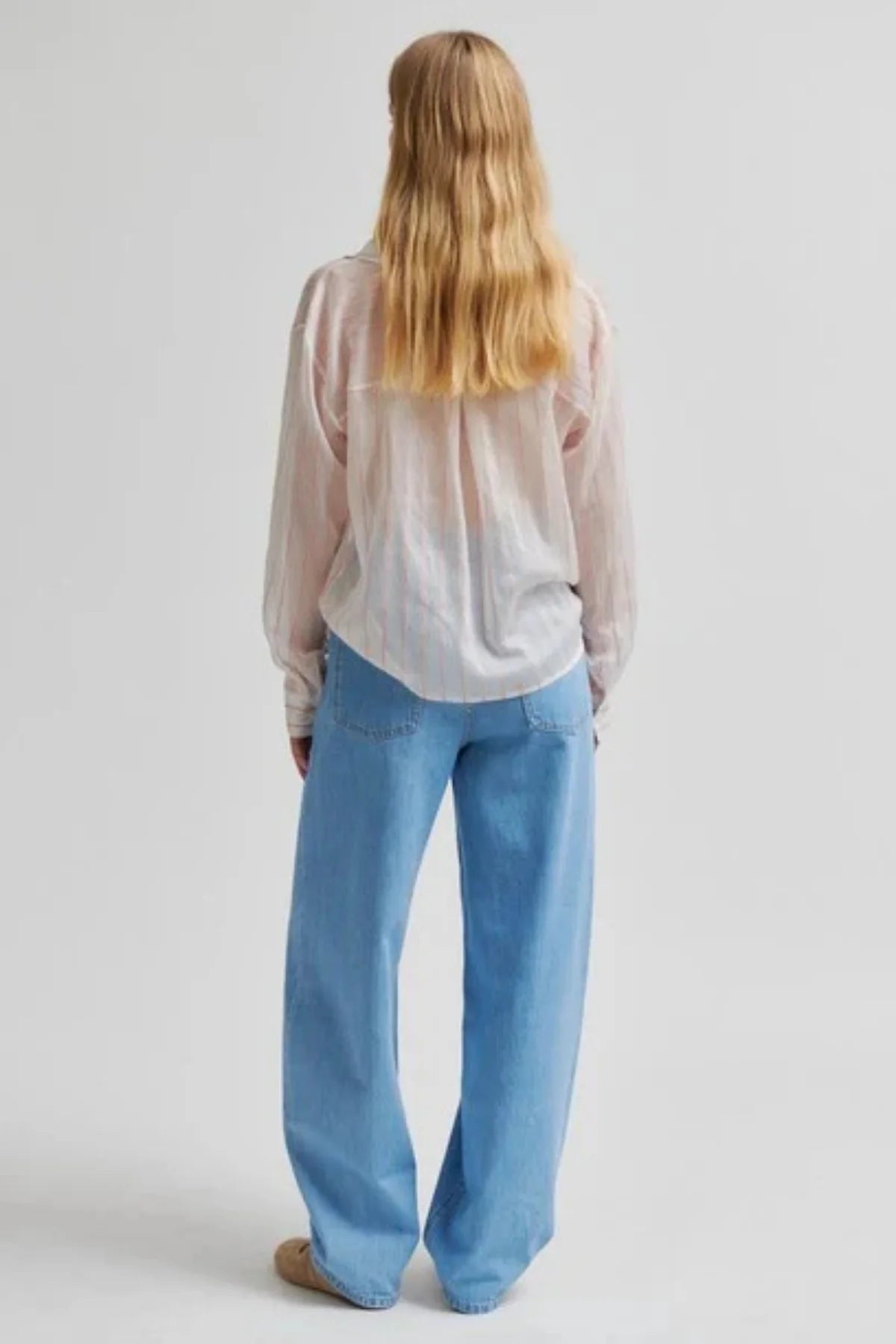 A woman with long blonde hair is shown from behind wearing a sheer, light pink long-sleeve top and light blue wide-leg jeans against a plain light gray background.