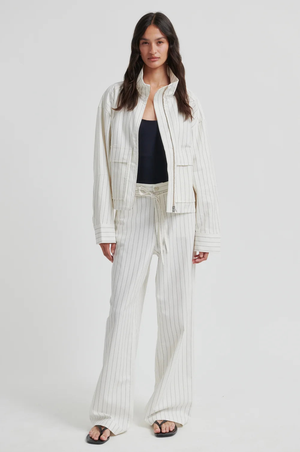 A model stands against a light studio background wearing a matching cream-colored pinstriped jacket and wide-leg trousers set, styled with a black tank top and black sandals.