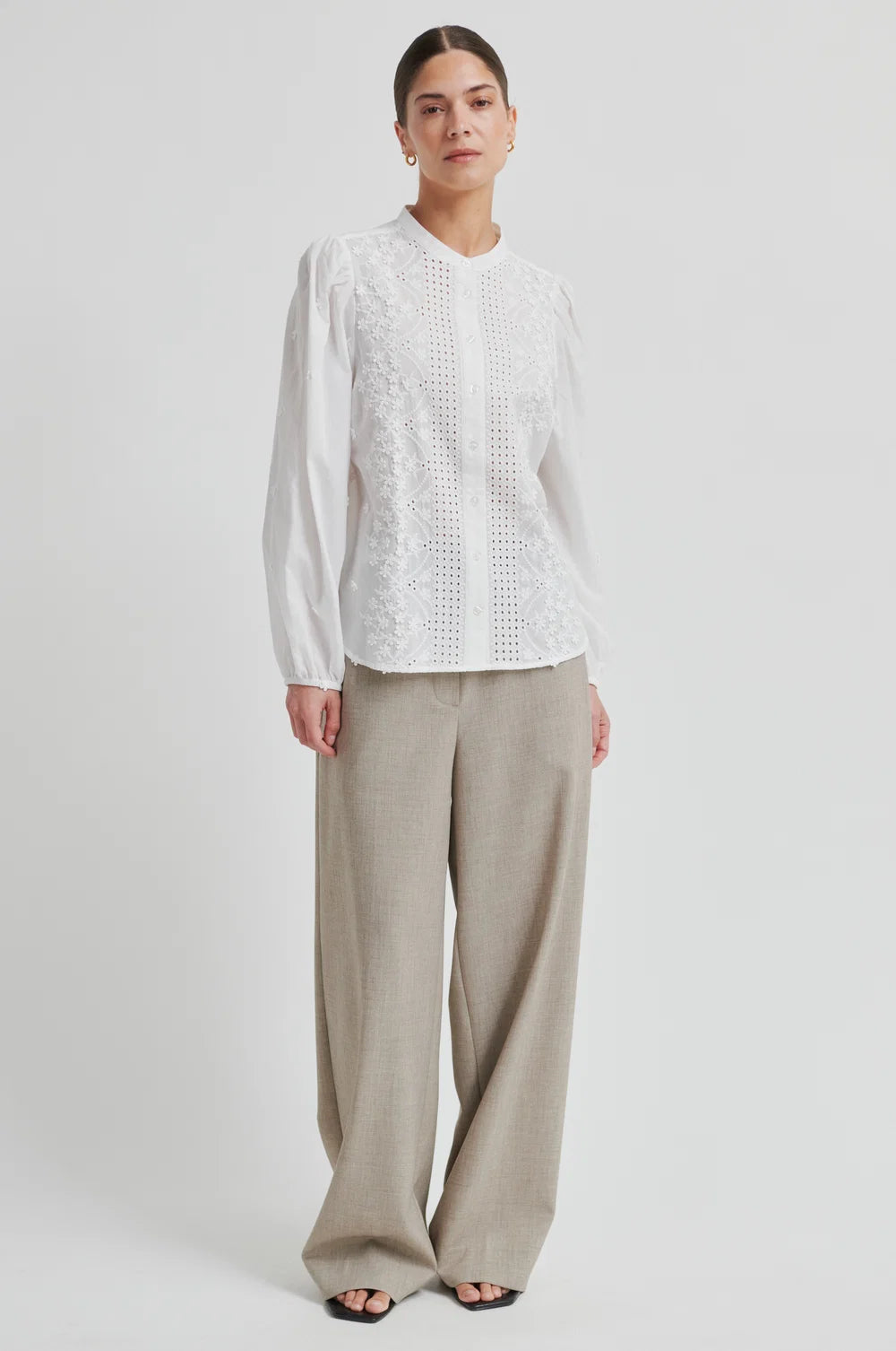 Woman wearing a white blouse with intricate patterns and beige pants  on a plain background