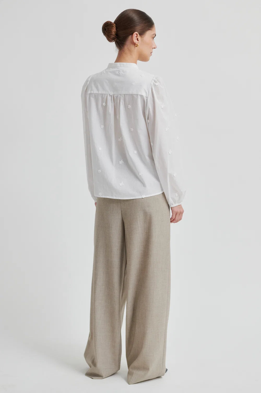 Woman shown from the back wearing  a white blouse with intricate patterns and beige pants  on a plain background