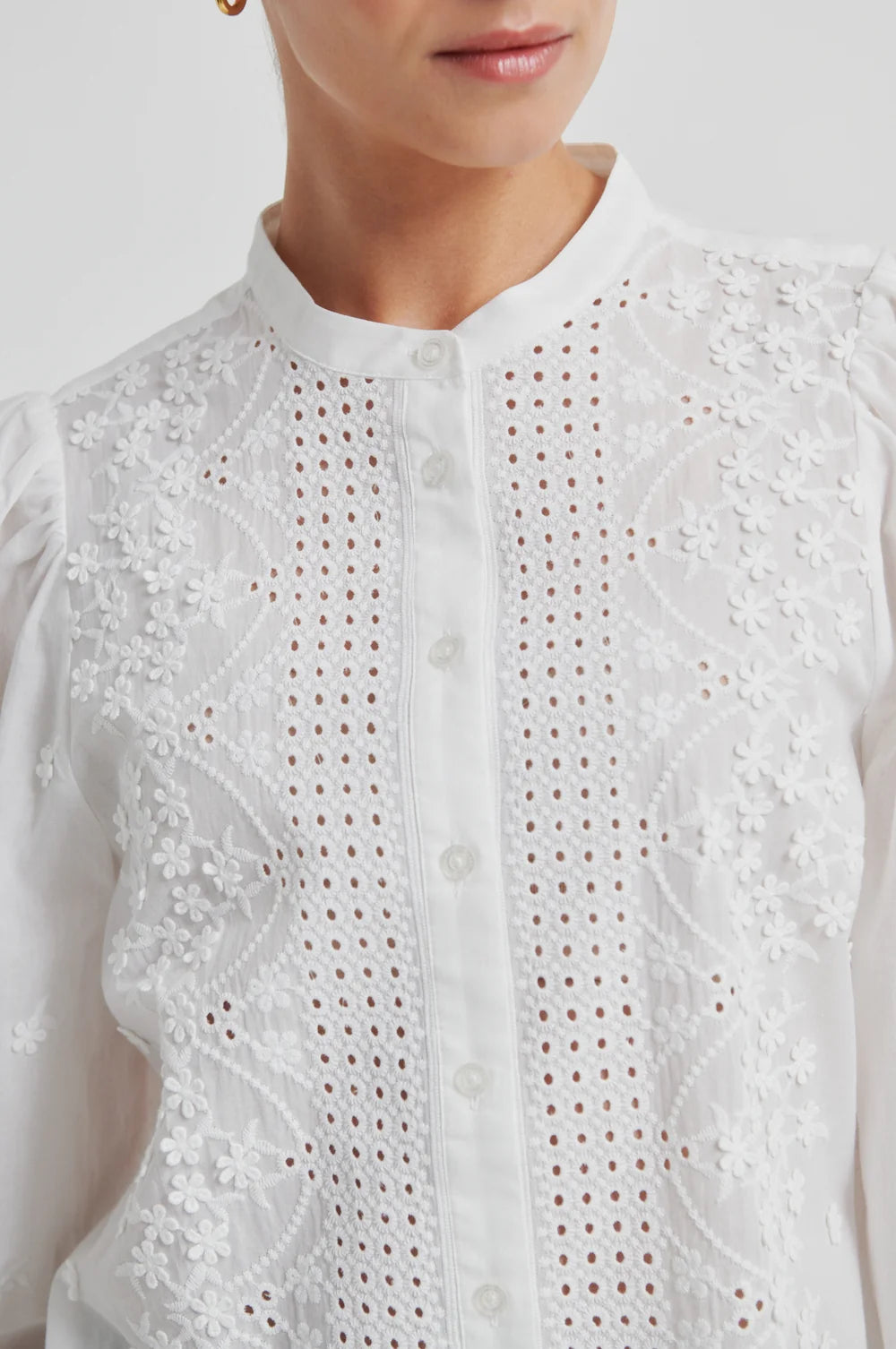Woman up close wearing a white blouse with intricate patternd  on a plain background