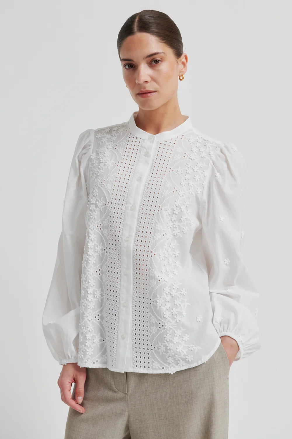 Woman wearing a white blouse with intricate patterns and beige pants  on a plain background