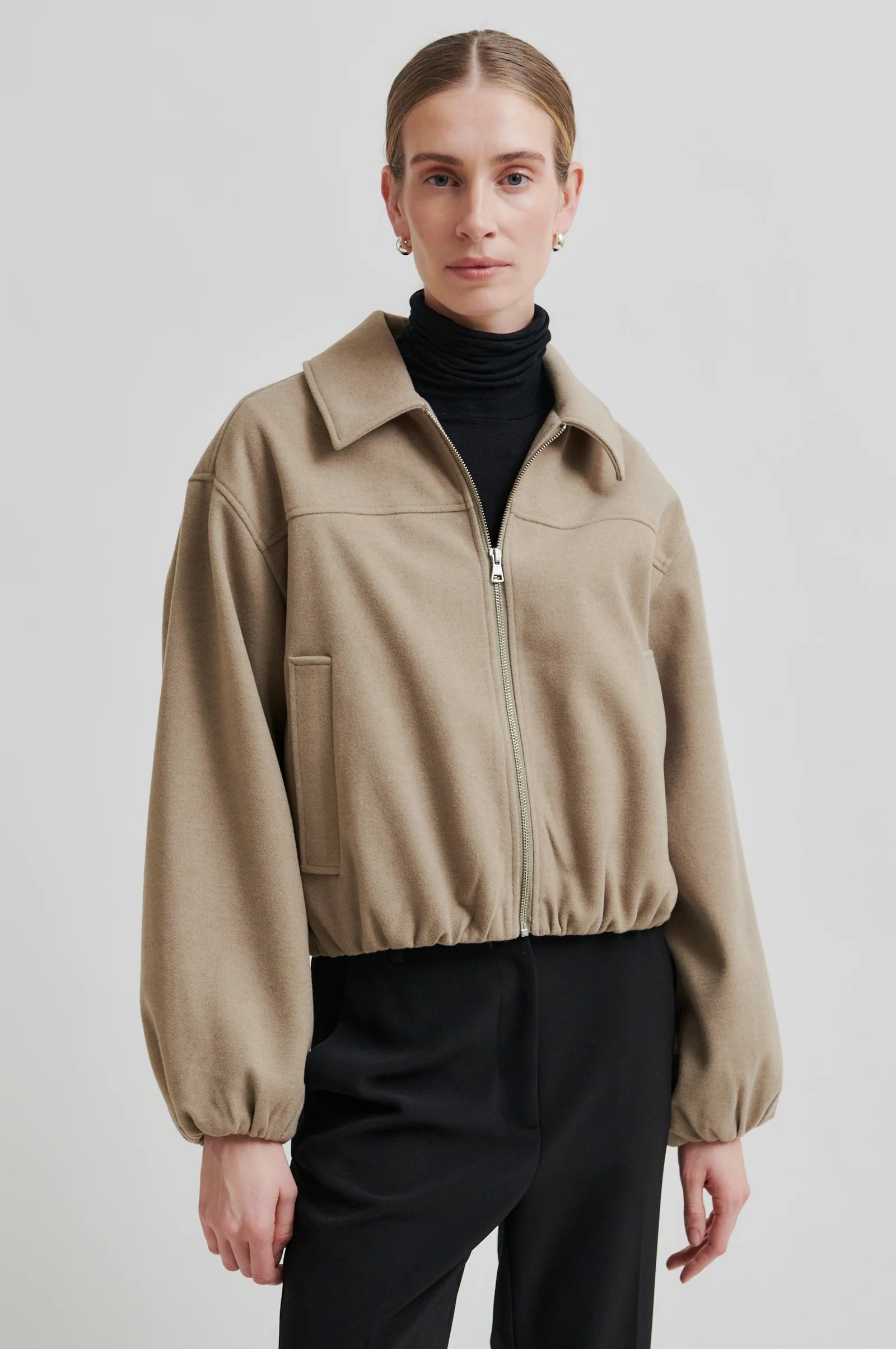 Person wearing a beige jacket over a black turtleneck and pants on a light gray background