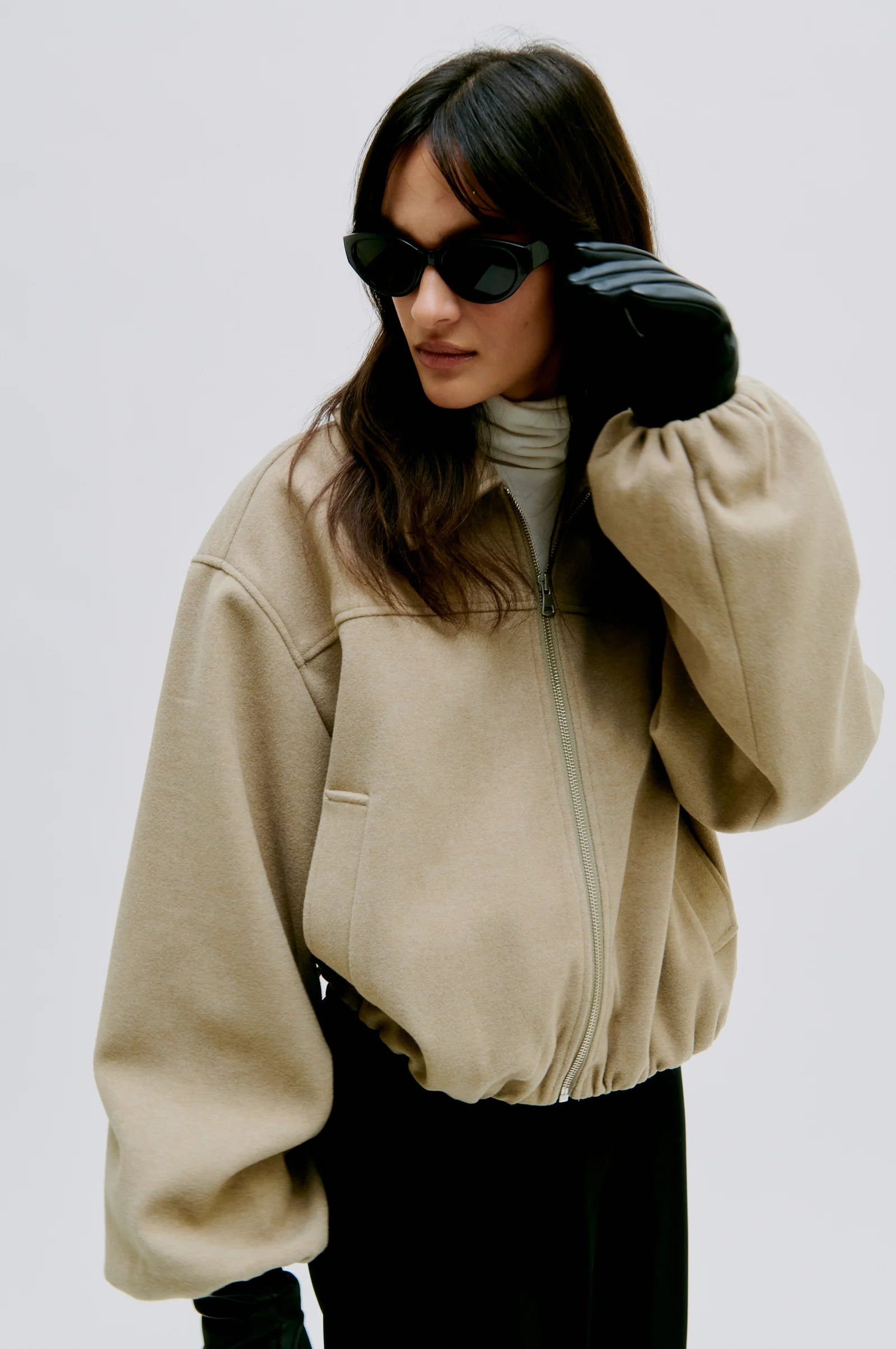 Person wearing a beige jacket and black pants, black gloves and sunglasses on a plain background