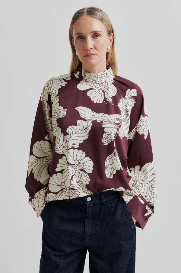 Woman wearing a burgundy blouse with floral pattern and dark pants on a light gray background