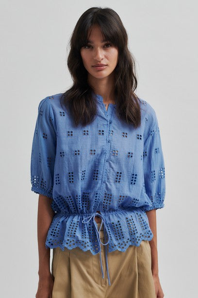 Model wearing blue broderie short sleeve shirt with tan shorts on a light background. 