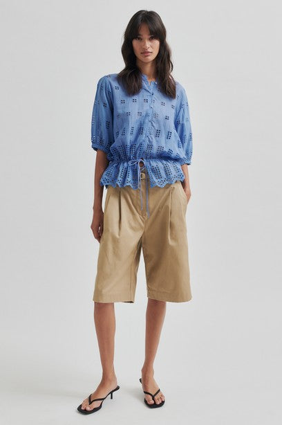 Model wearing blue broderie short sleeve shirt with tan shorts on a light background. 