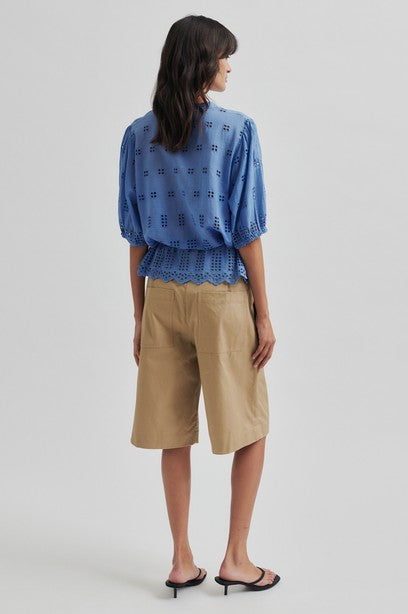Model wearing blue broderie shirt with tan shorts on a light background. 