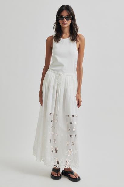 Woman wearing a white sleeveless top and white skirt with cut-out details on a plain background