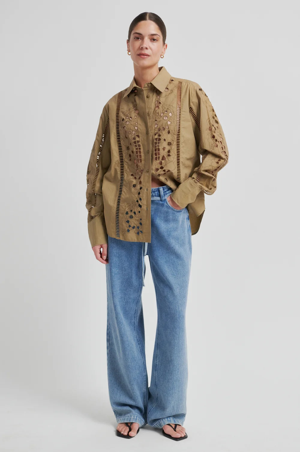 A model stands against a light gray studio background wearing a tan button-up shirt with dark embroidered details, paired with wide-leg light blue jeans and black sandals.