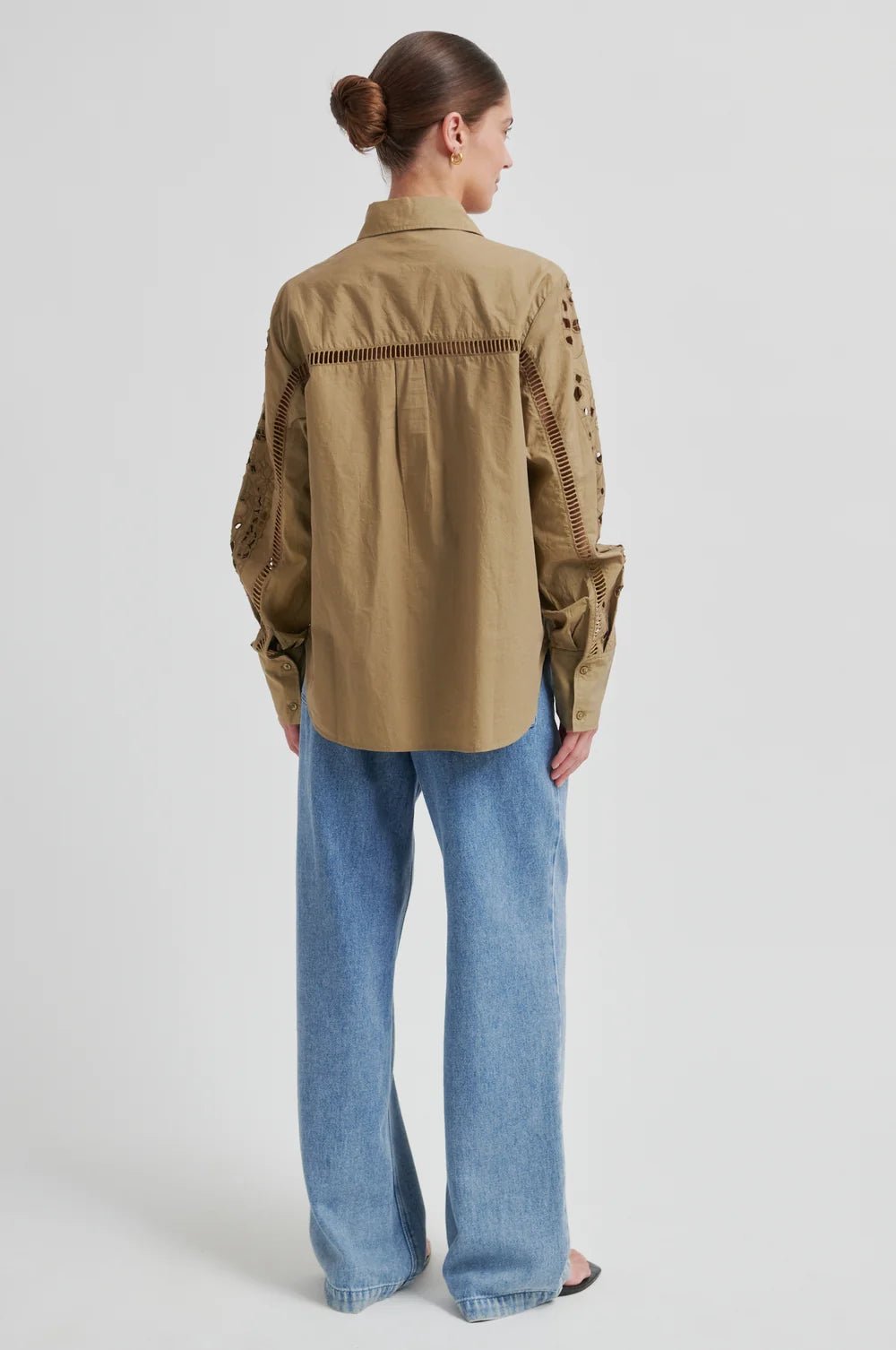 From the back a model wearing a beige button-up shirt with intricate laser-cutout embroidery and vertical stitched detailing on the chest and sleeves, set against a plain light gray background.