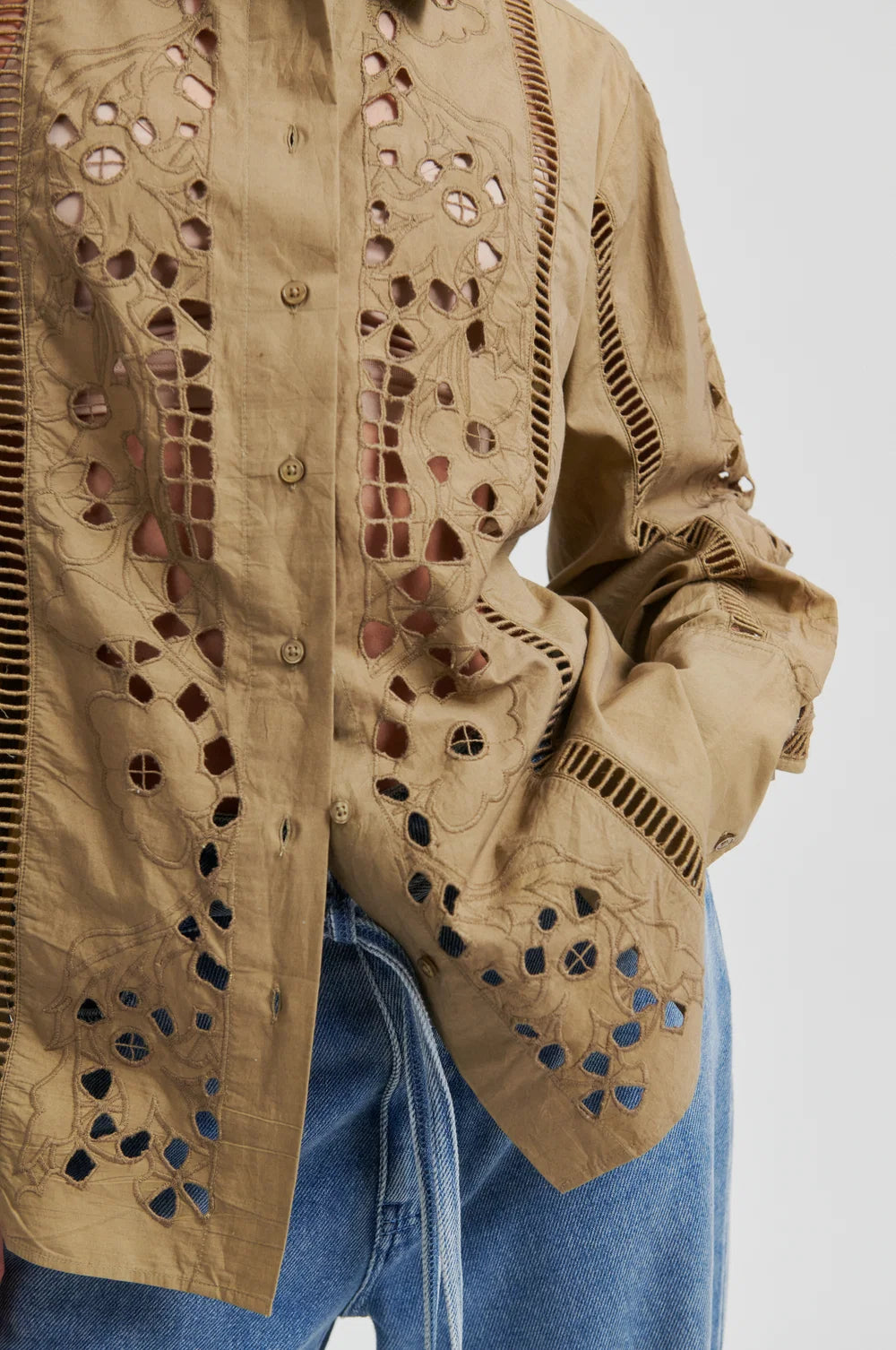 A close-up of a model wearing a beige button-up shirt with intricate laser-cutout embroidery and vertical stitched detailing on the chest and sleeves, set against a plain light gray background.