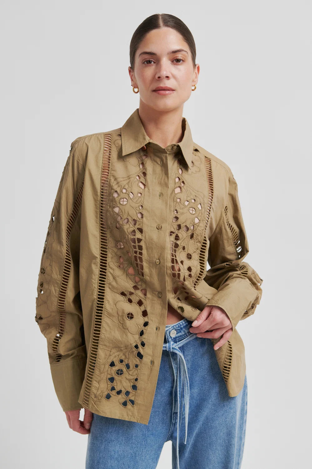 A close-up of a model wearing a beige button-up shirt with intricate laser-cutout embroidery and vertical stitched detailing on the chest and sleeves, set against a plain light gray background.