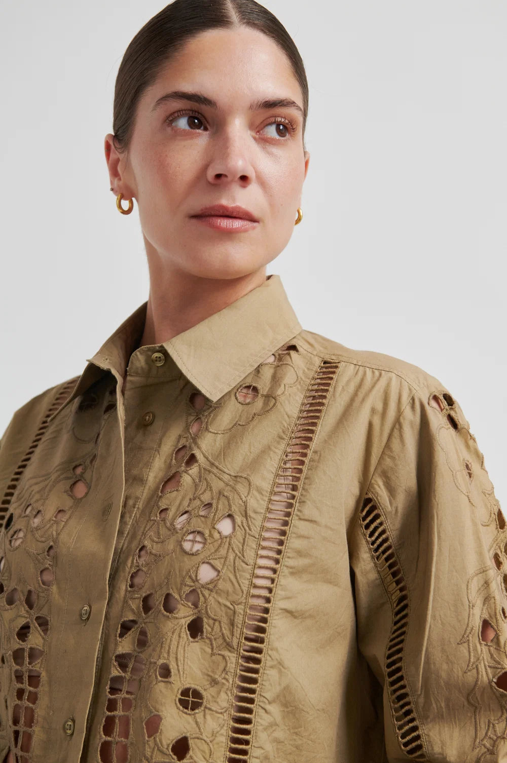 A close-up of a model wearing a beige button-up shirt with intricate laser-cutout embroidery and vertical stitched detailing on the chest and sleeves, set against a plain light gray background.