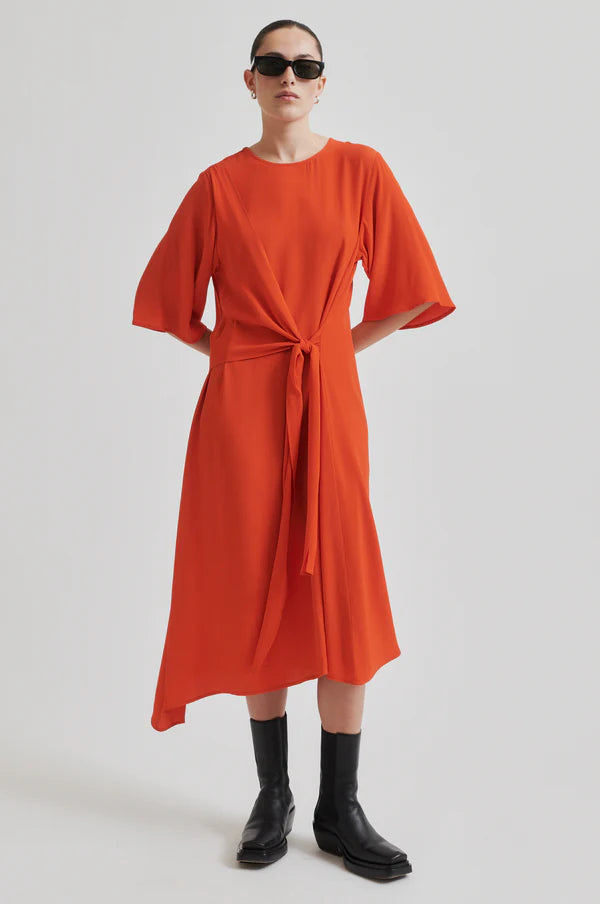Model wearing orange short sleeve midi dress with asymmetrical hem and tie waist. Wearing black boots and sunglasses. 