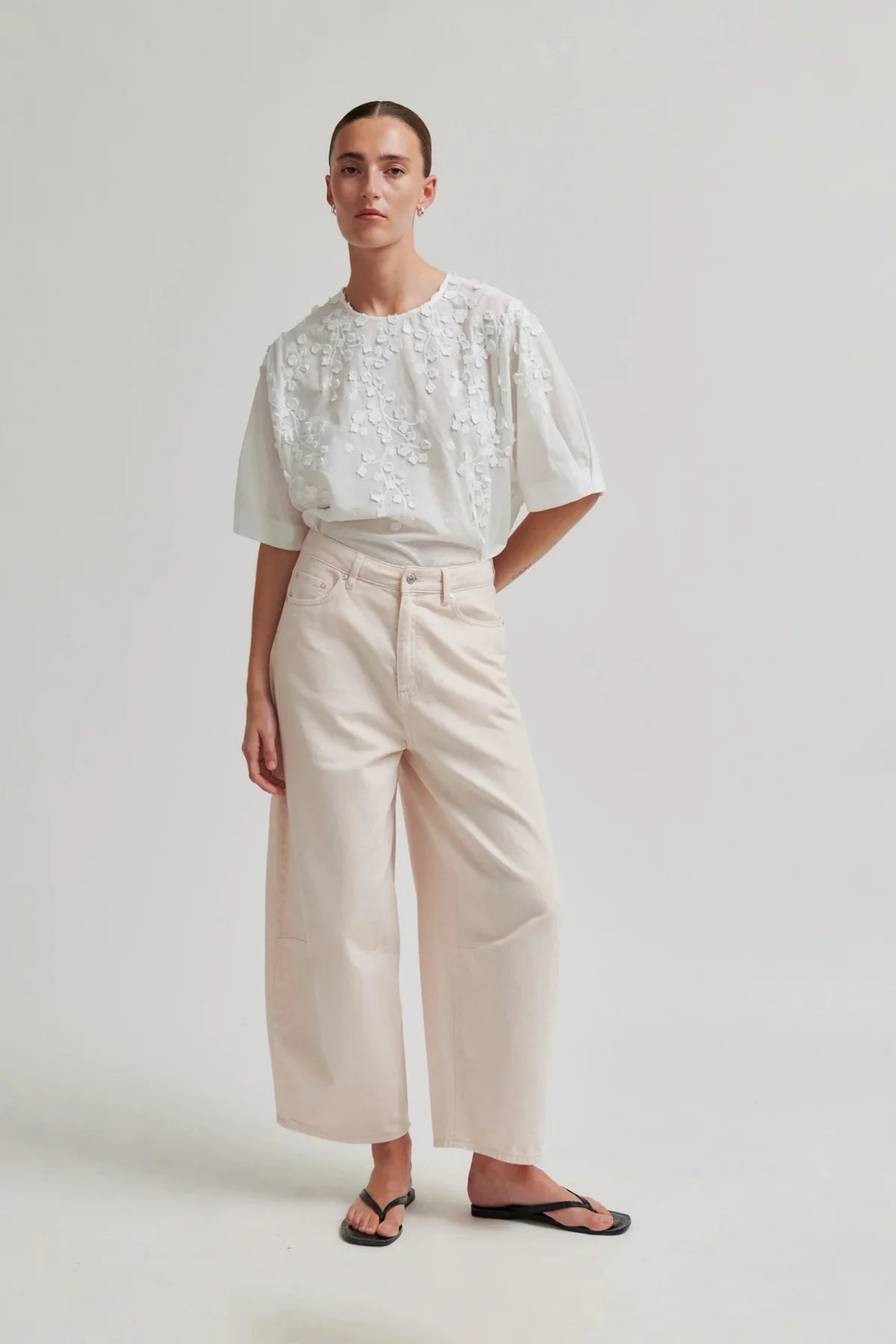 A model stands against a light grey studio background wearing a white short-sleeve top with floral embroidery and cream wide-leg cropped pants, paired with black flip-flop sandals.