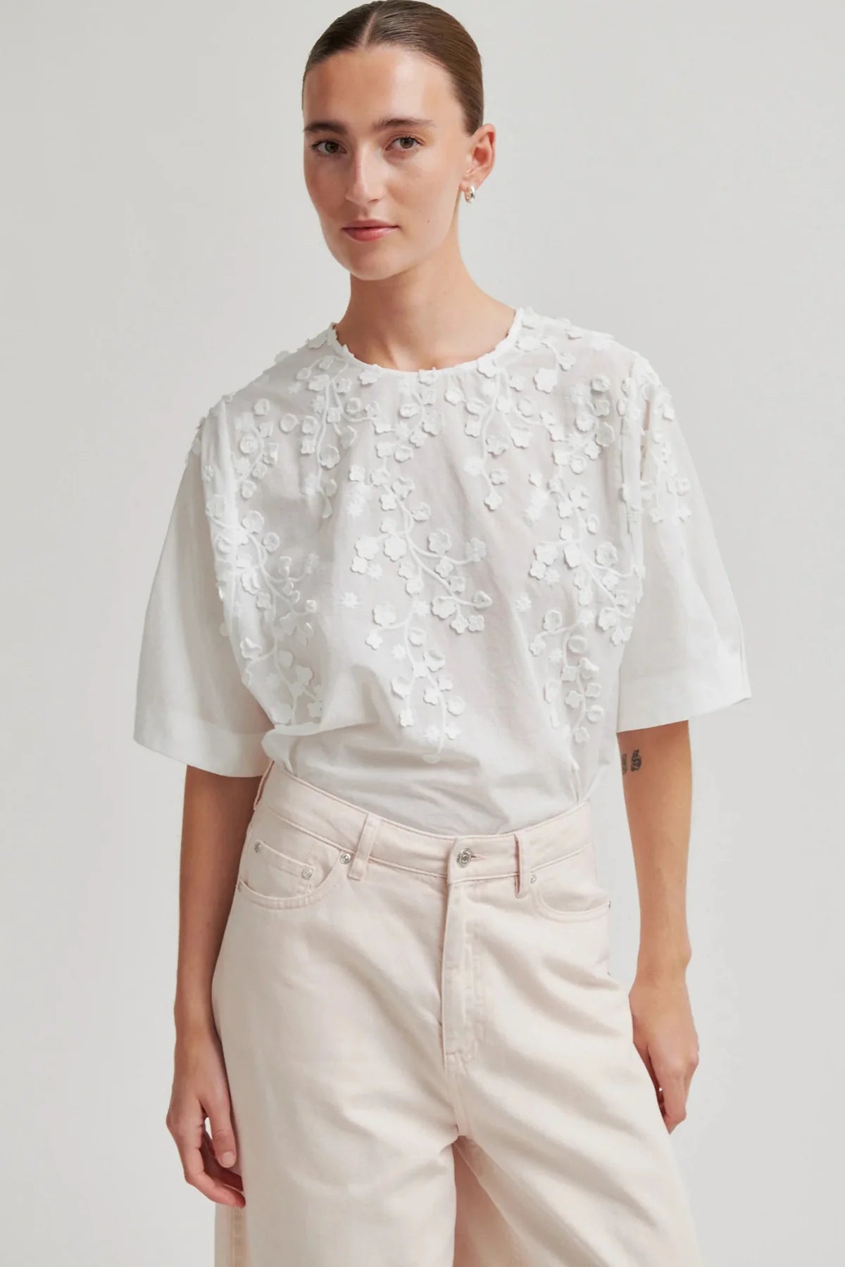 A woman modeling a white short-sleeve blouse with 3D floral embroidery on the bodice, paired with light beige trousers, against a plain light gray studio background.