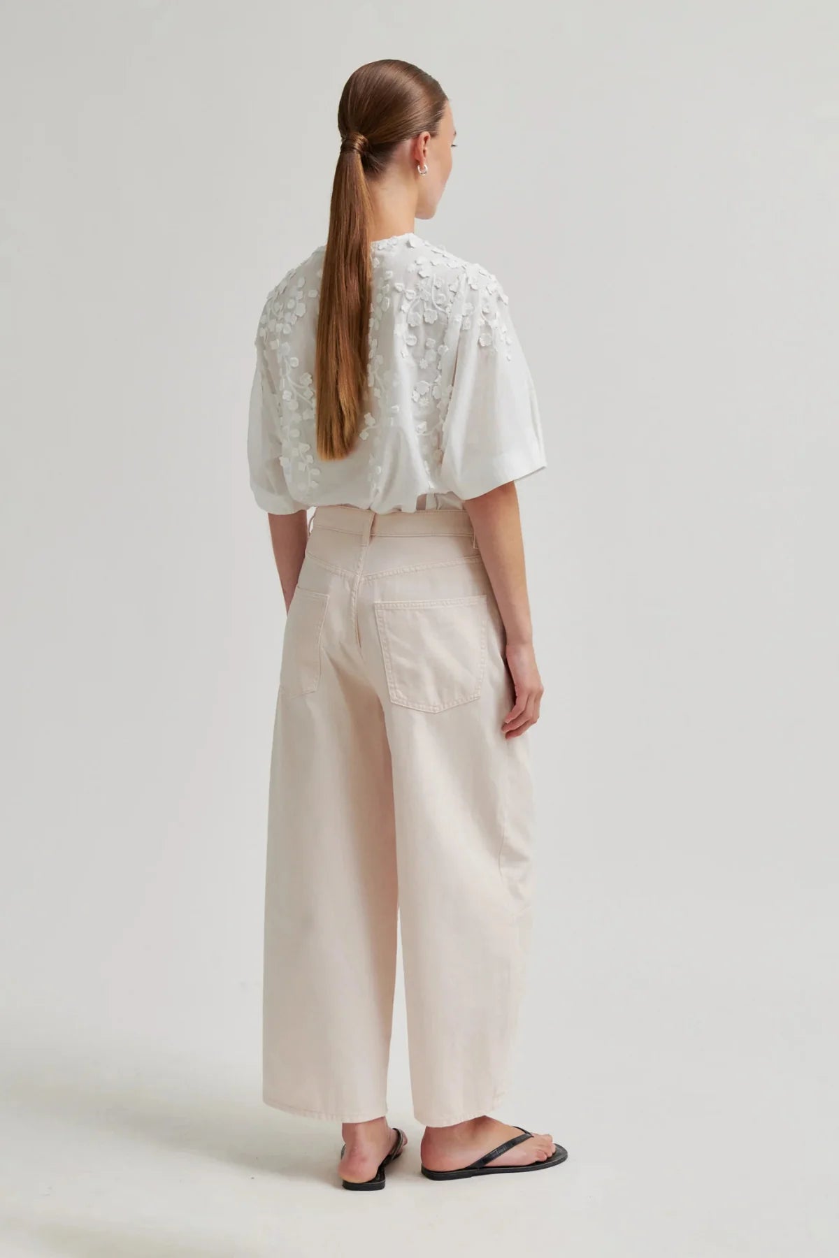 A woman shown from the back wearing a white short-sleeve blouse with floral lace detailing on the shoulders and high-waisted, wide-leg cream trousers, paired with black flip-flop sandals, against a plain light gray studio background.