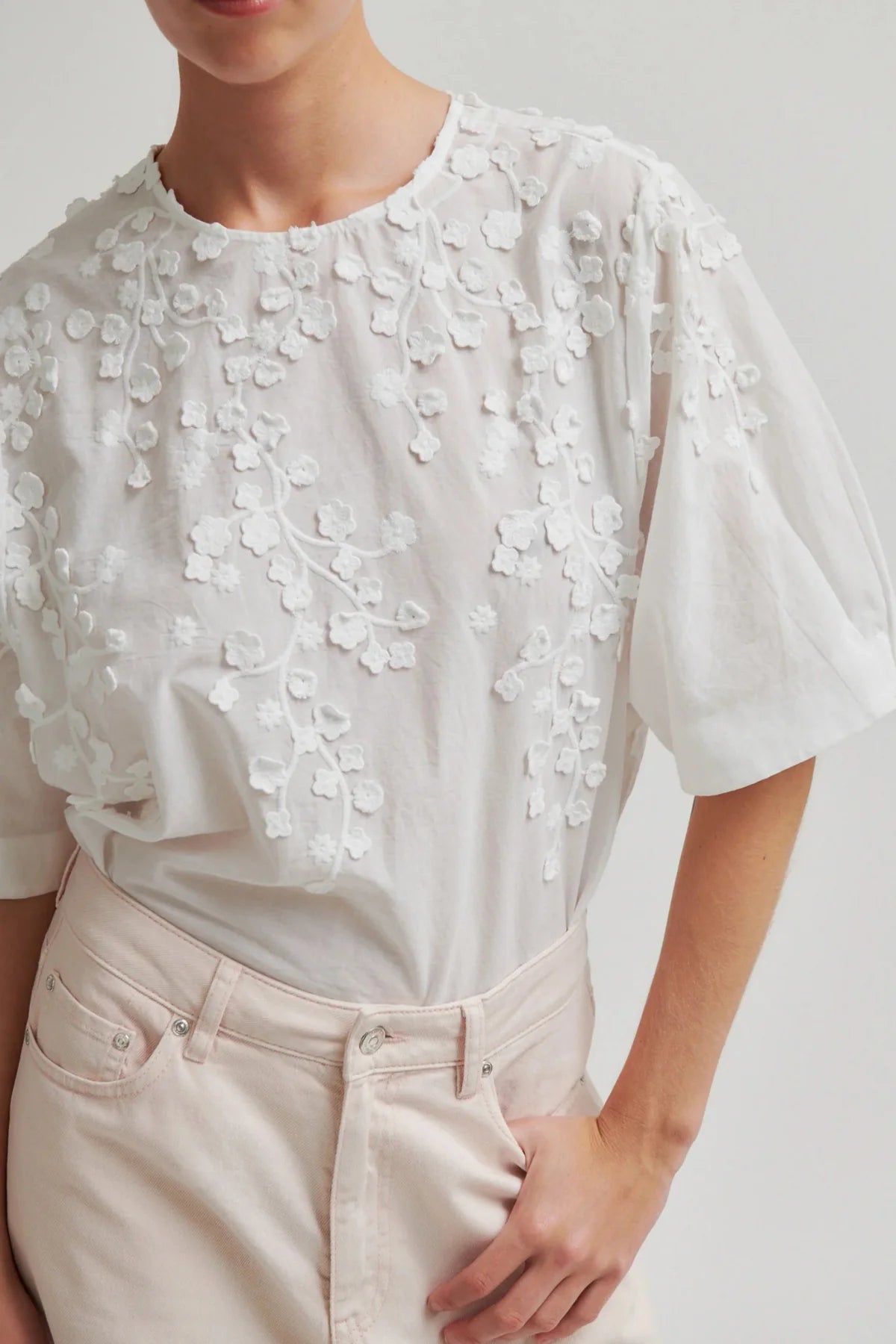 A close-up of a person wearing a white short-sleeve blouse with intricate 3D floral embroidery and applique across the chest, paired with light beige trousers.