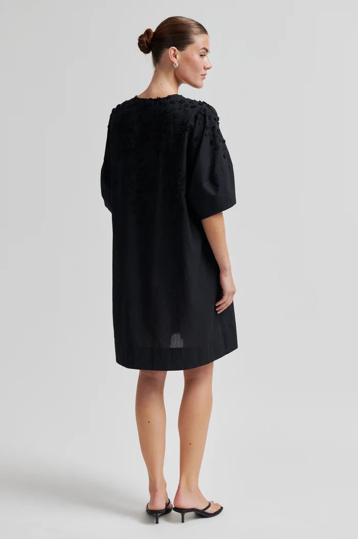Woman shown from the back wearing a black midi dress  with embroidery on the front and shoulder against a grey background. 
