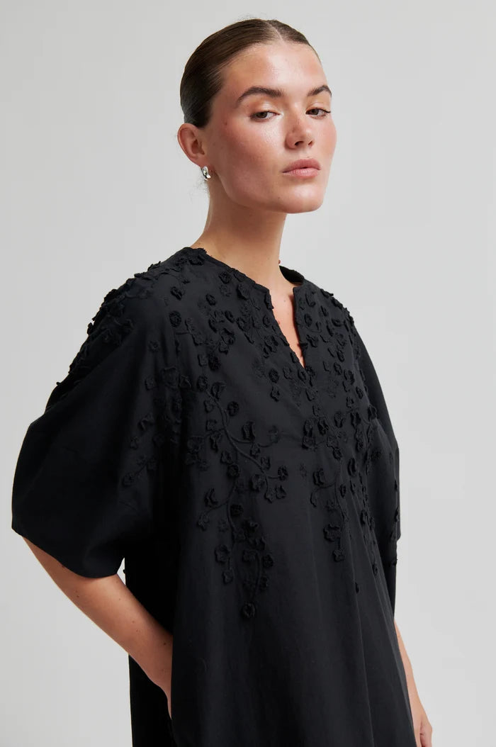 Woman shown side on wearing a black midi dress with floral embroidery on a light grey background