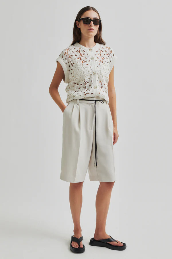 Model wearing white crochet top and white pleated shorts with a black drawstring cord belt on a light grey background