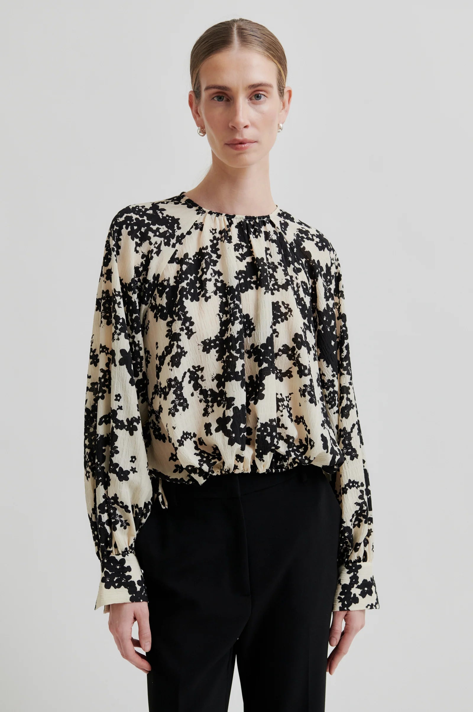 Women wearing a long sleeve cream blouse with black floral print and black pants on a white background