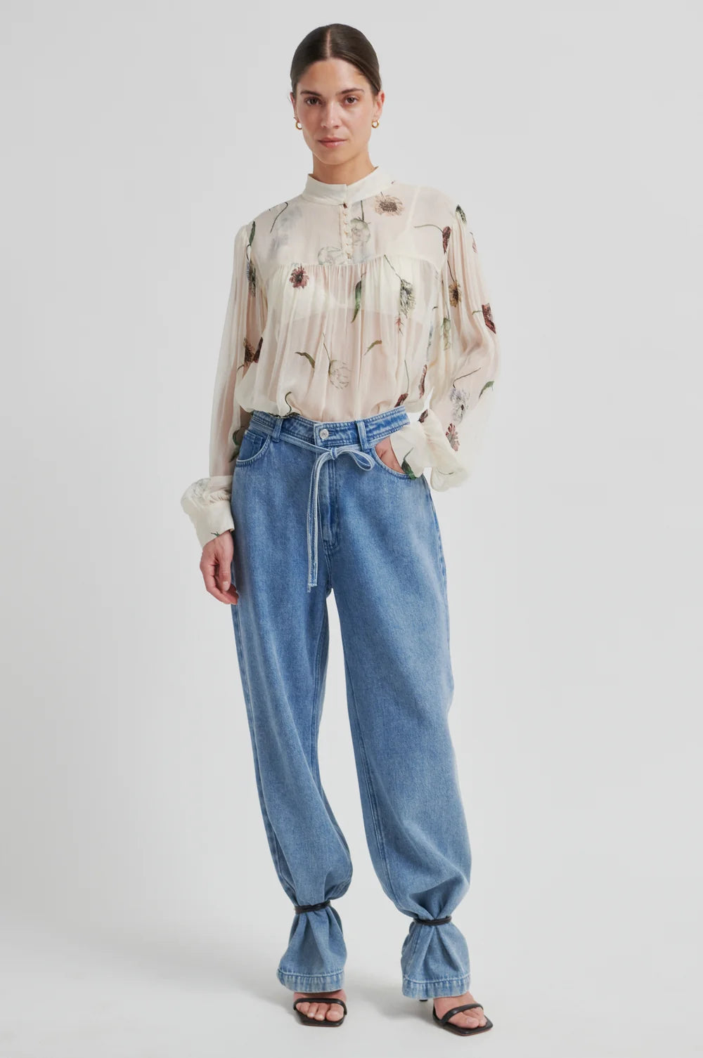 Model  wearing a floral-patterned blouse  and denim pants a neutral background 