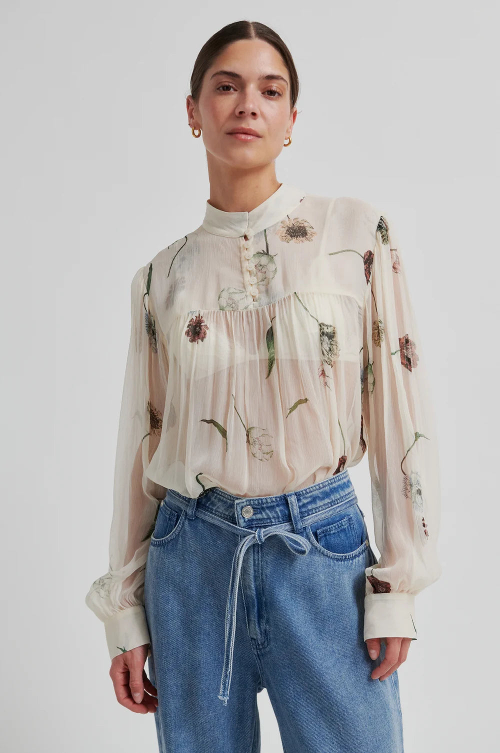 Model  wearing a floral-patterned blouse  and denim pants a neutral background 