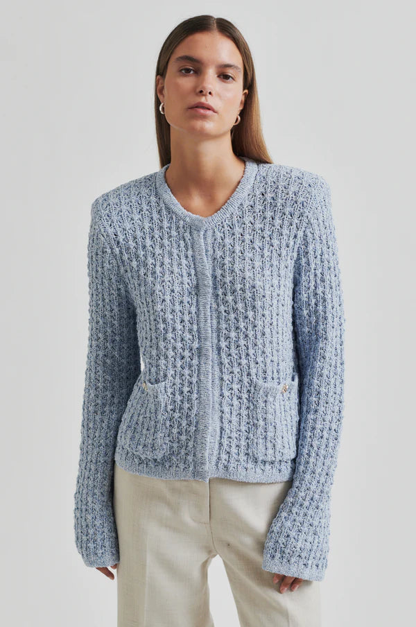 Model wearing a blue knitted cardigan and beige pants up close to see cardigan detail. 