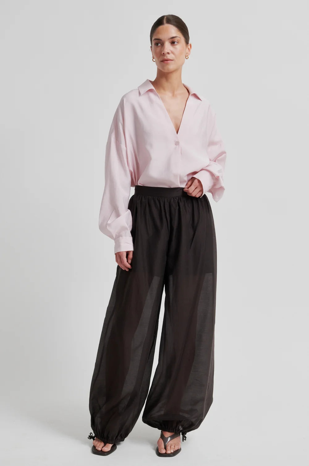 Model wearing chocolate brown wide-leg pants  that taper and a a pink shirt on a white background