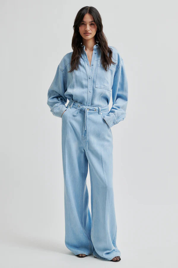 Model wearing light blue denim jean in drapy fabric with waist tie. Paired with light blue denim shirt in same fabric. 
