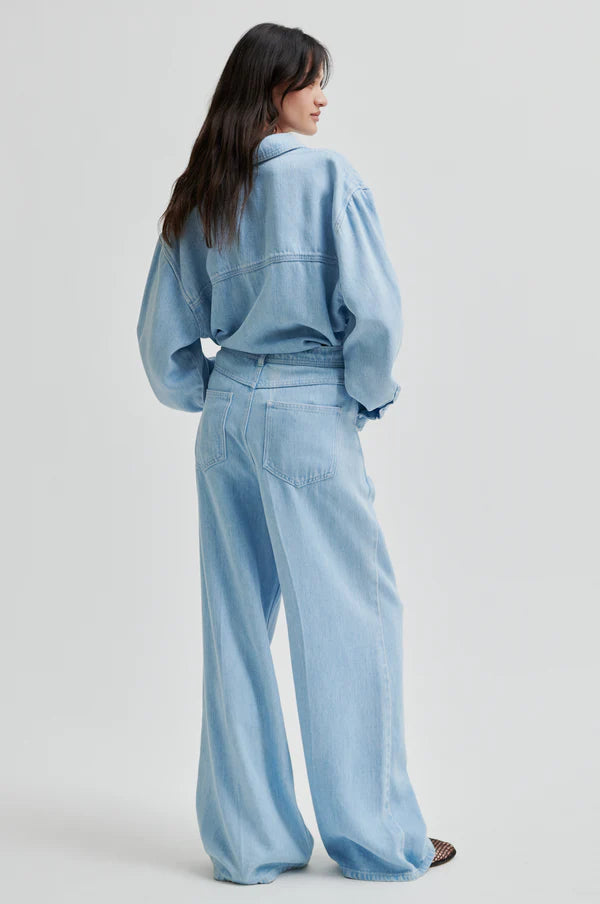 Model wearing light blue denim jean in drapy fabric with waist tie. Paired with light blue denim shirt in same fabric. 
