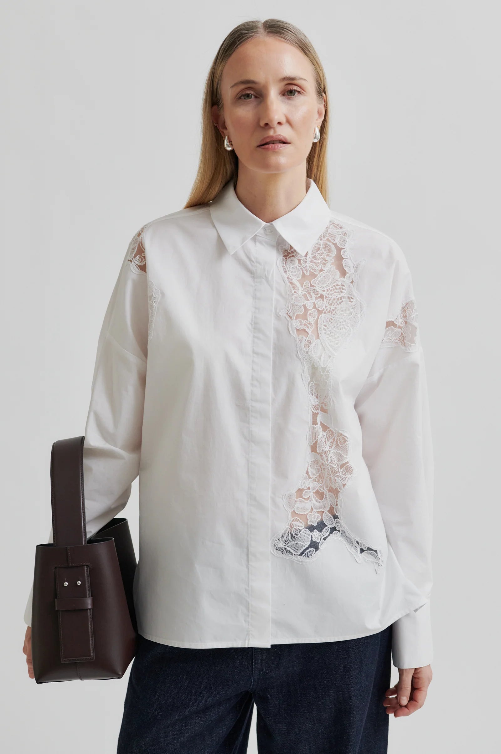 Woman wearing a white shirt with lace detailing and dark pants holding a brown leather bag on a light grey background