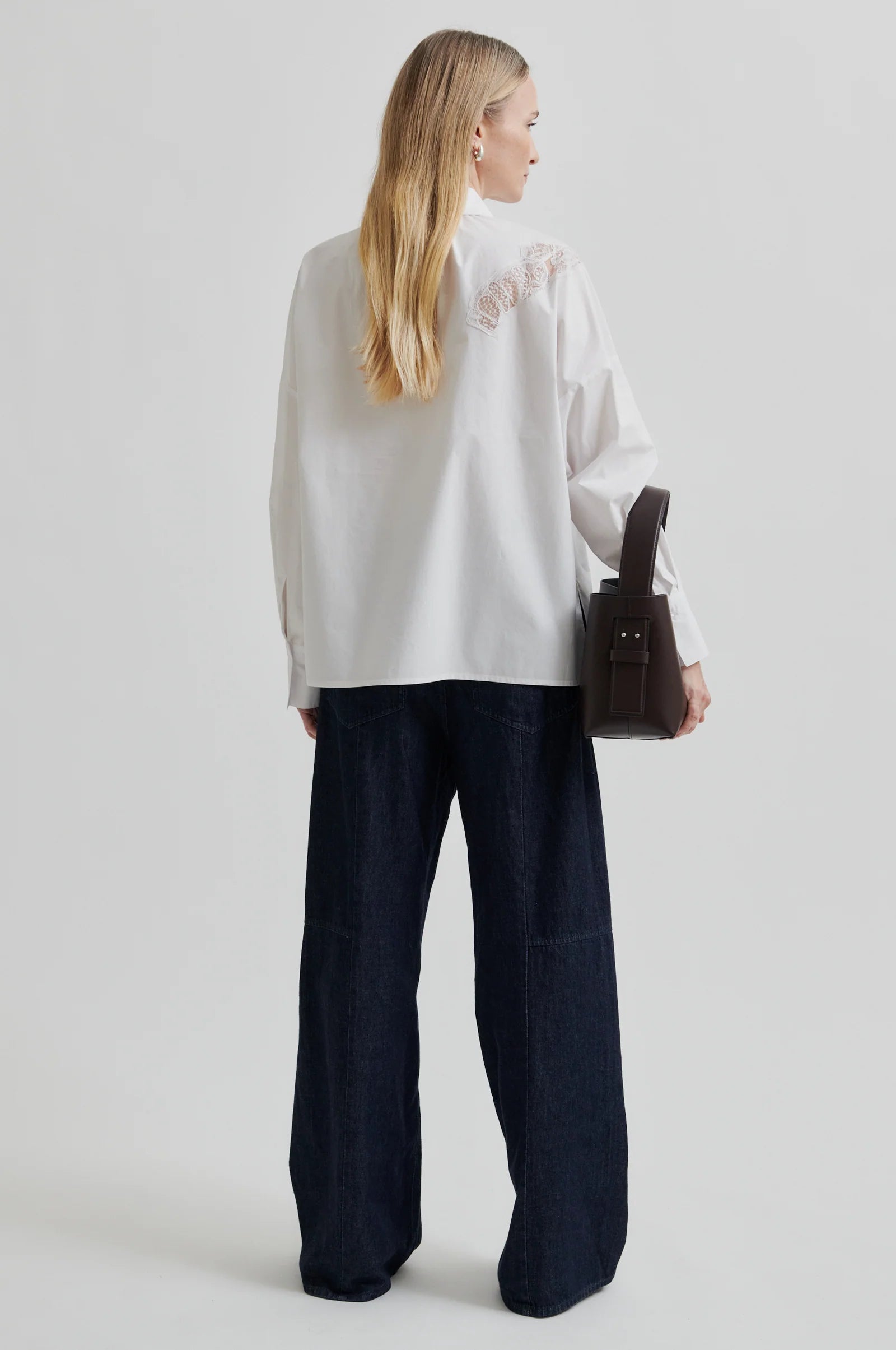 Woman wearing a white shirt with lace detailing and dark pants holding a brown leather bag on a light grey background