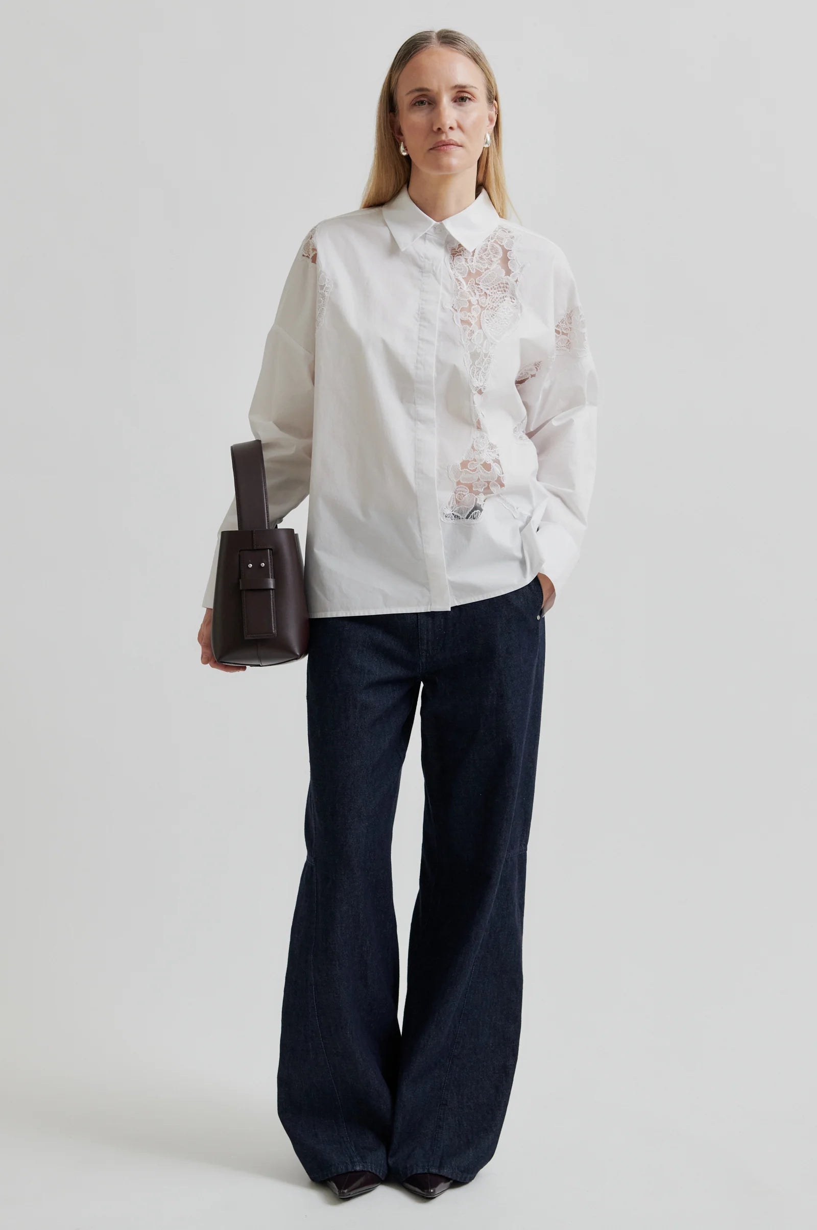 Woman wearing a white shirt with lace detailing and dark pants holding a brown leather bag on a light grey background