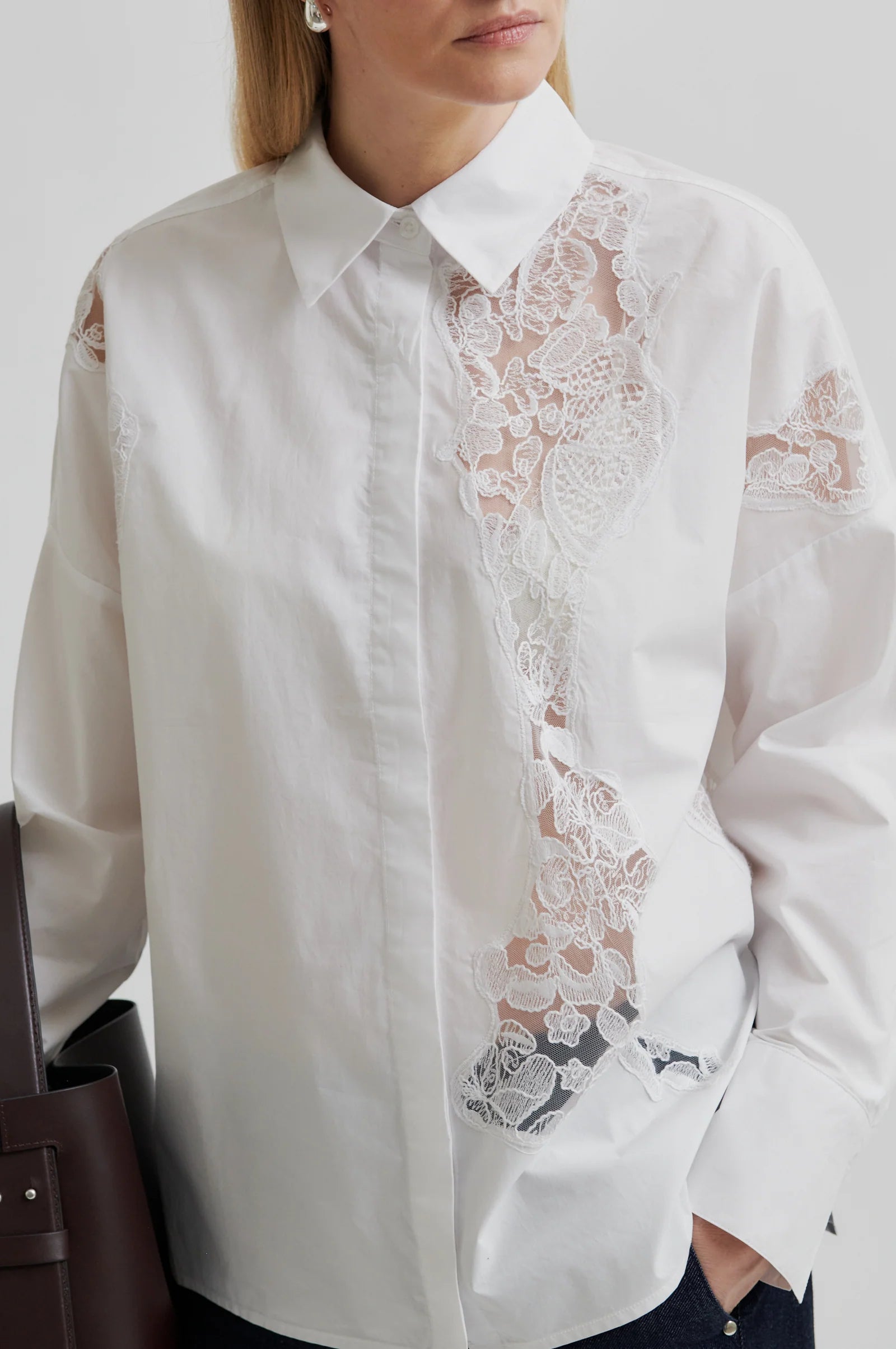 Woman wearing a white shirt with lace detailing and dark pants holding a brown leather bag on a light grey background