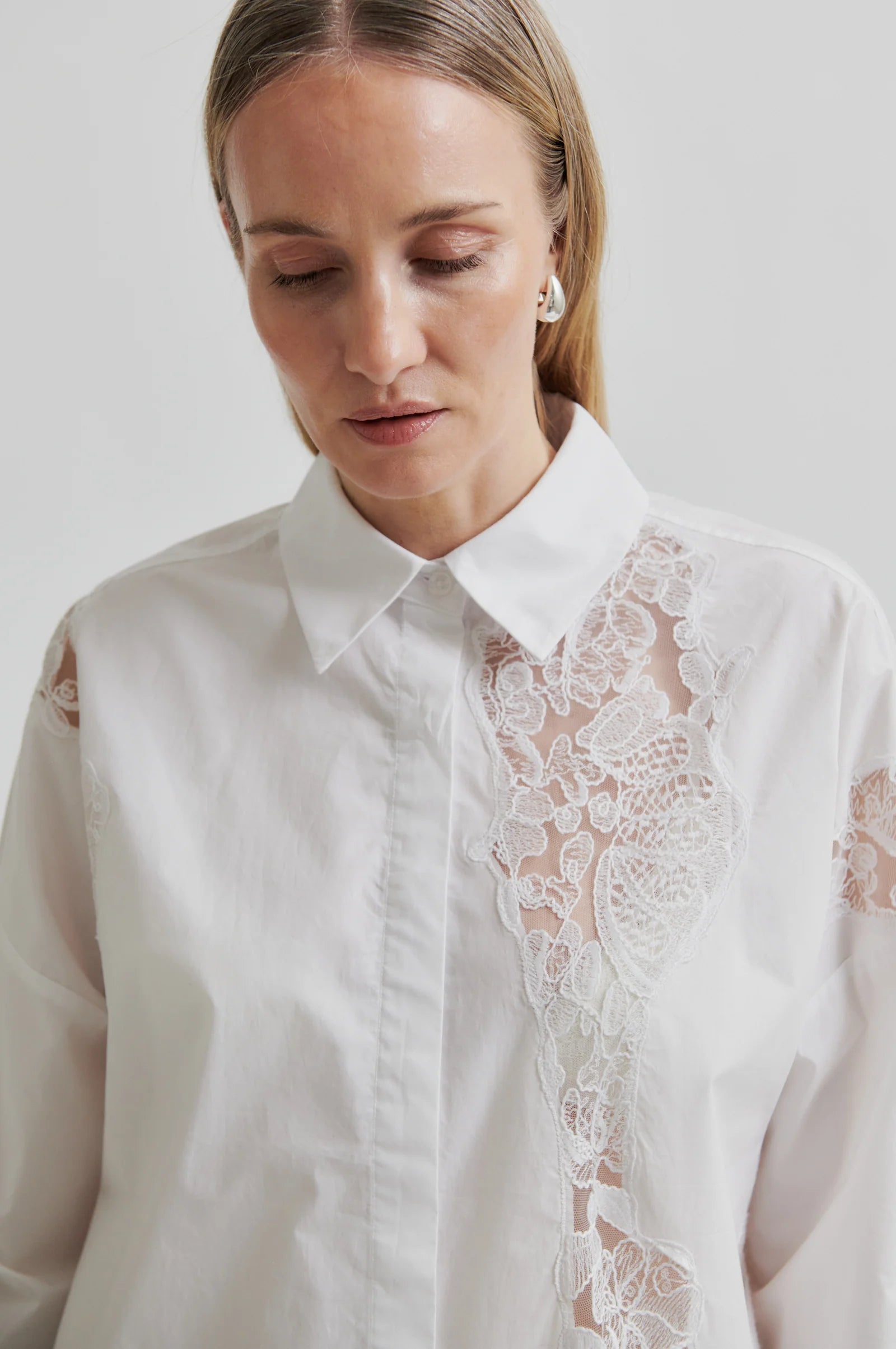 Woman wearing a white shirt with lace detailing and dark pants holding a brown leather bag on a light grey background
