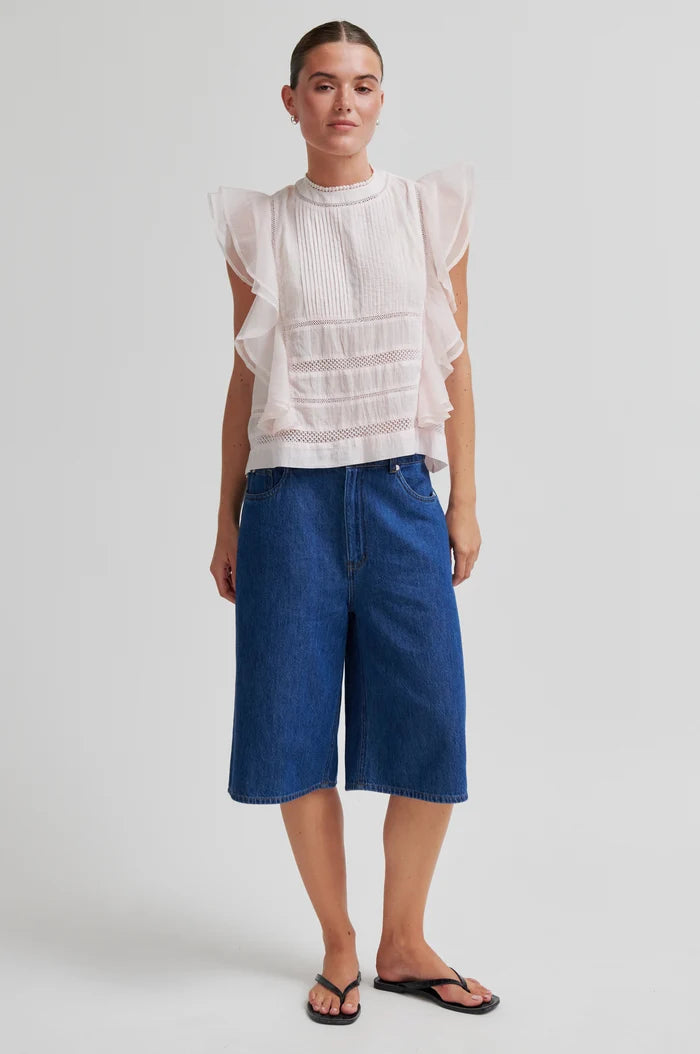 A model stands against a light gray studio background wearing a white ruffled sleeveless blouse, blue denim cropped jeans, and black flip-flops.