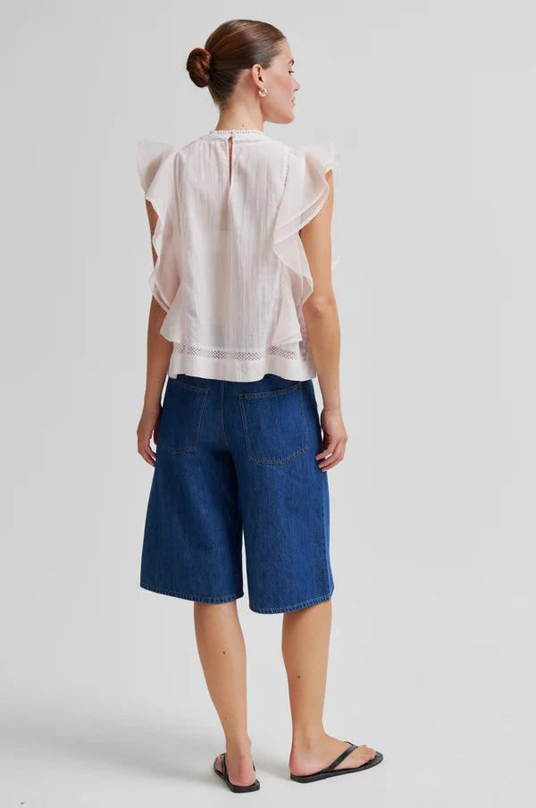 Woman shown from the back wearing a white blouse with ruffle sleeveless detail and blue denim shorts on a white background