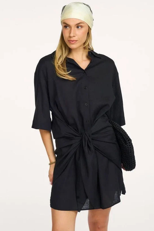 A woman modeling a black button-down shirt dress with three-quarter sleeves and a knotted waist, styled with a cream headband and a black woven clutch against a plain white background.