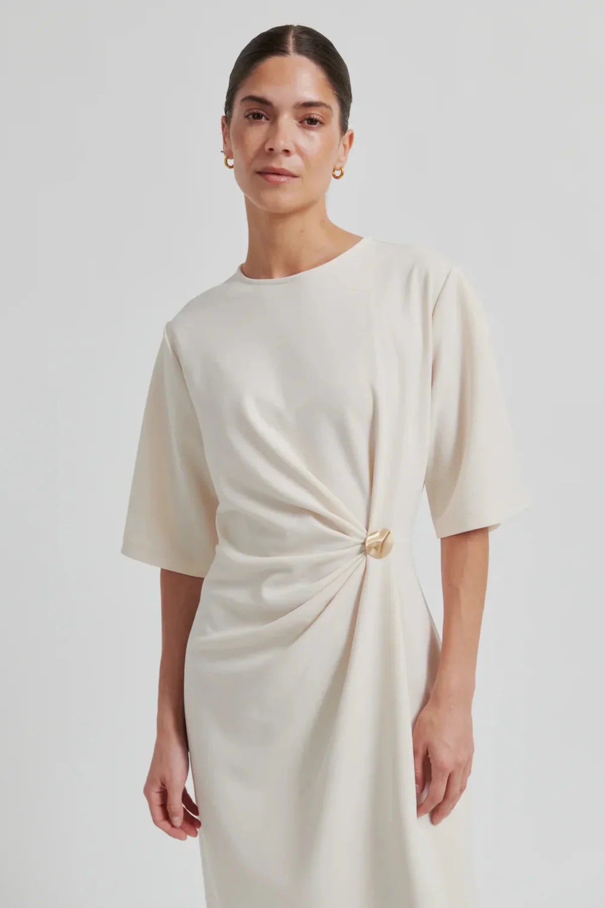 Woman wearing a beige dress with a knot detail and gold fastening on a plain background
