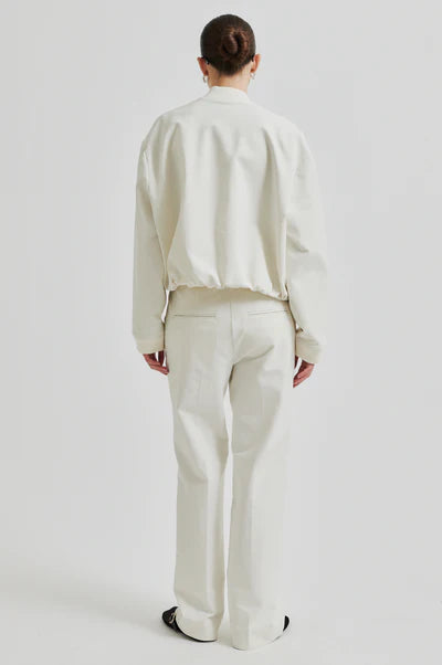 Model wearing drapy cream bomber jacket with front pockets. Paired with cream pants and black sunglasses.  Back view. 