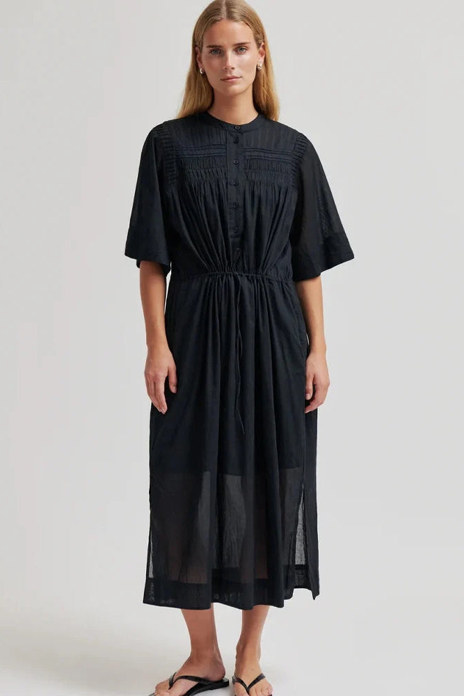 A woman modeling a black midi dress with short sleeves, a gathered waist, and a smocked neckline, shown against a plain light gray studio background.