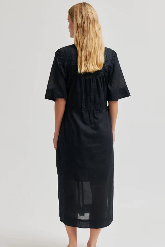 A woman viewed from the back wearing a black midi dress with short sleeves, a gathered waist, and sheer mesh panels at the hem, against a light gray studio background.