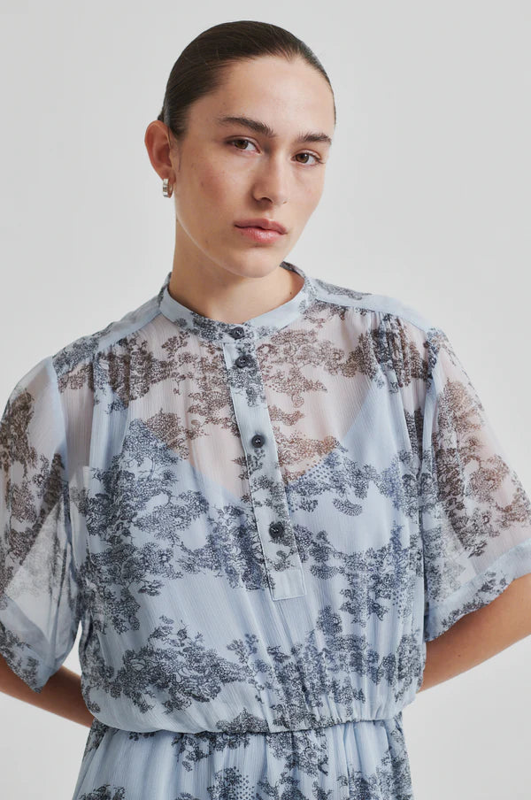 Model wearing a blue sheer floral dress with cap sleeve and print. 