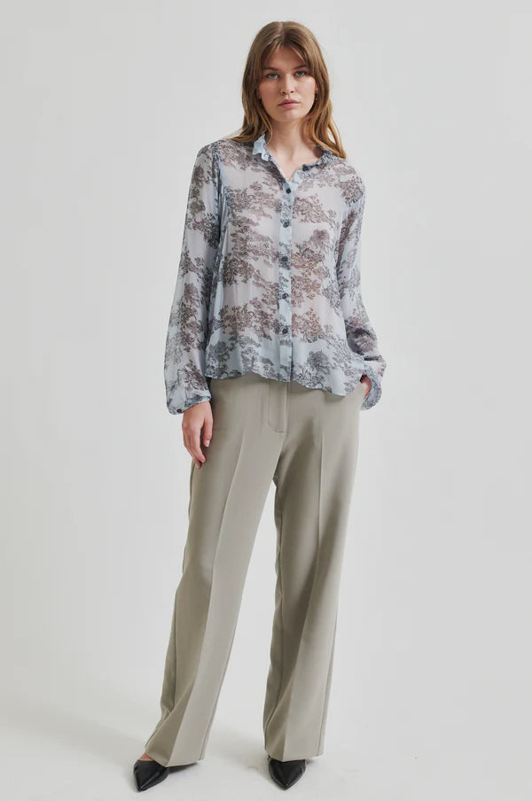 Model wearing soft blue sheer shirt with grey floral print paired with soft tan pants. 
