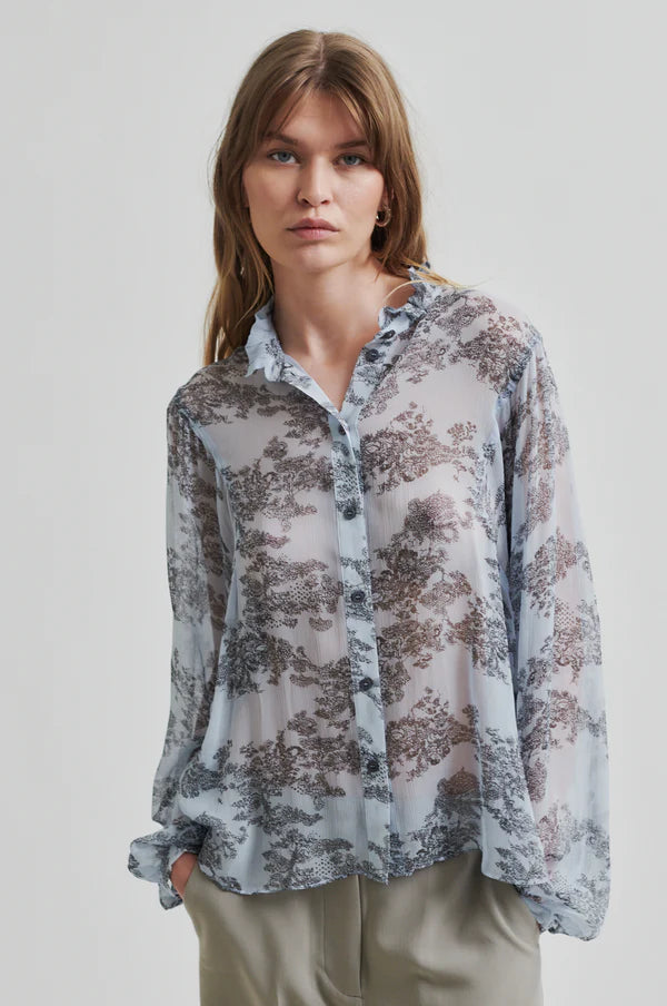Model wearing soft blue sheer shirt with grey floral print paired with soft tan pants. 