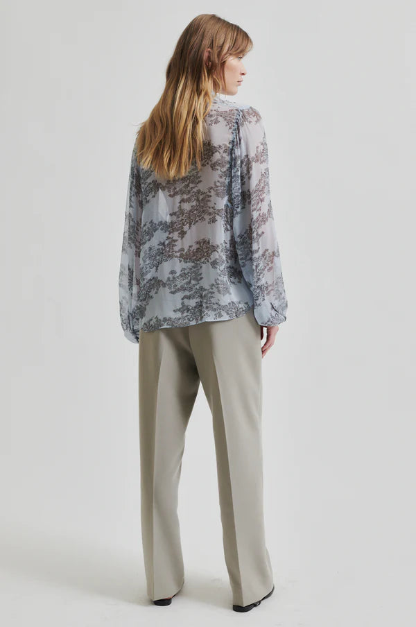 Model wearing soft blue sheer shirt with grey floral print paired with soft tan pants. 