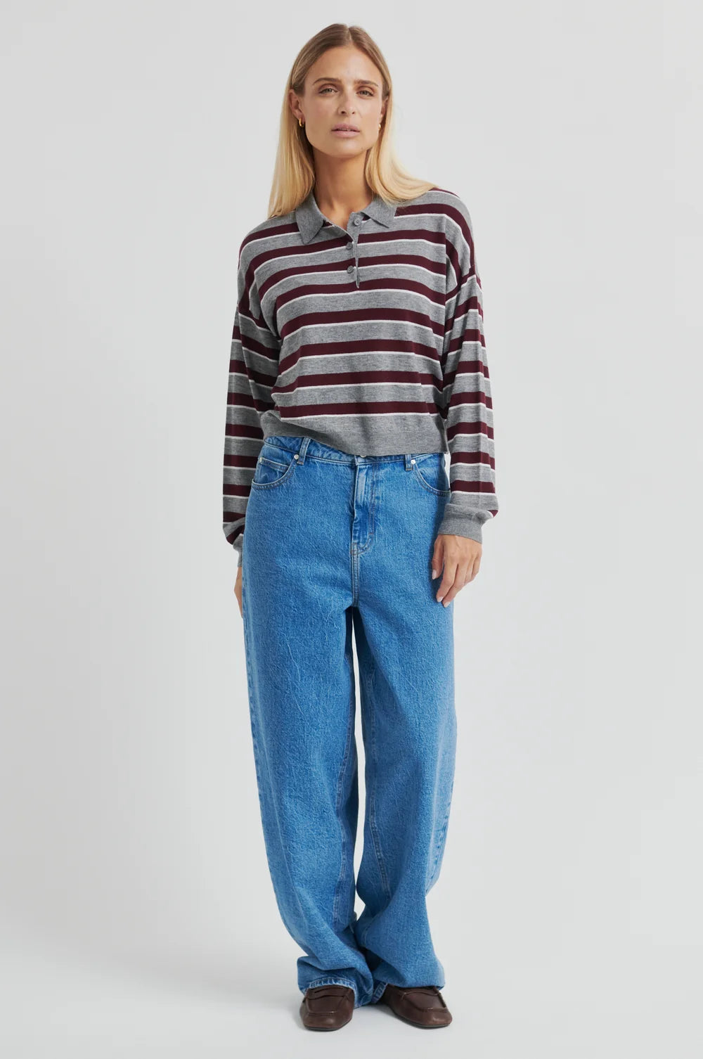 Women wearing a striped cropped polo sweater  in grey and burgundy with jeans on a plain background.  